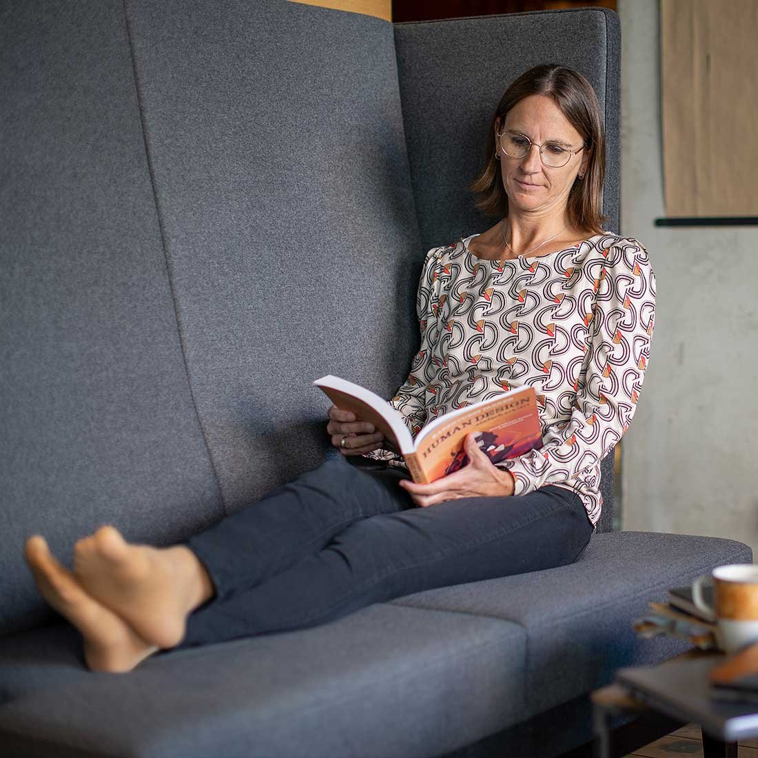 Performance Coach Manuela Hartmann lehnt auf einer grauen Eckbank und ist vertieft in Literatur zu Human Design für Führungskräfte